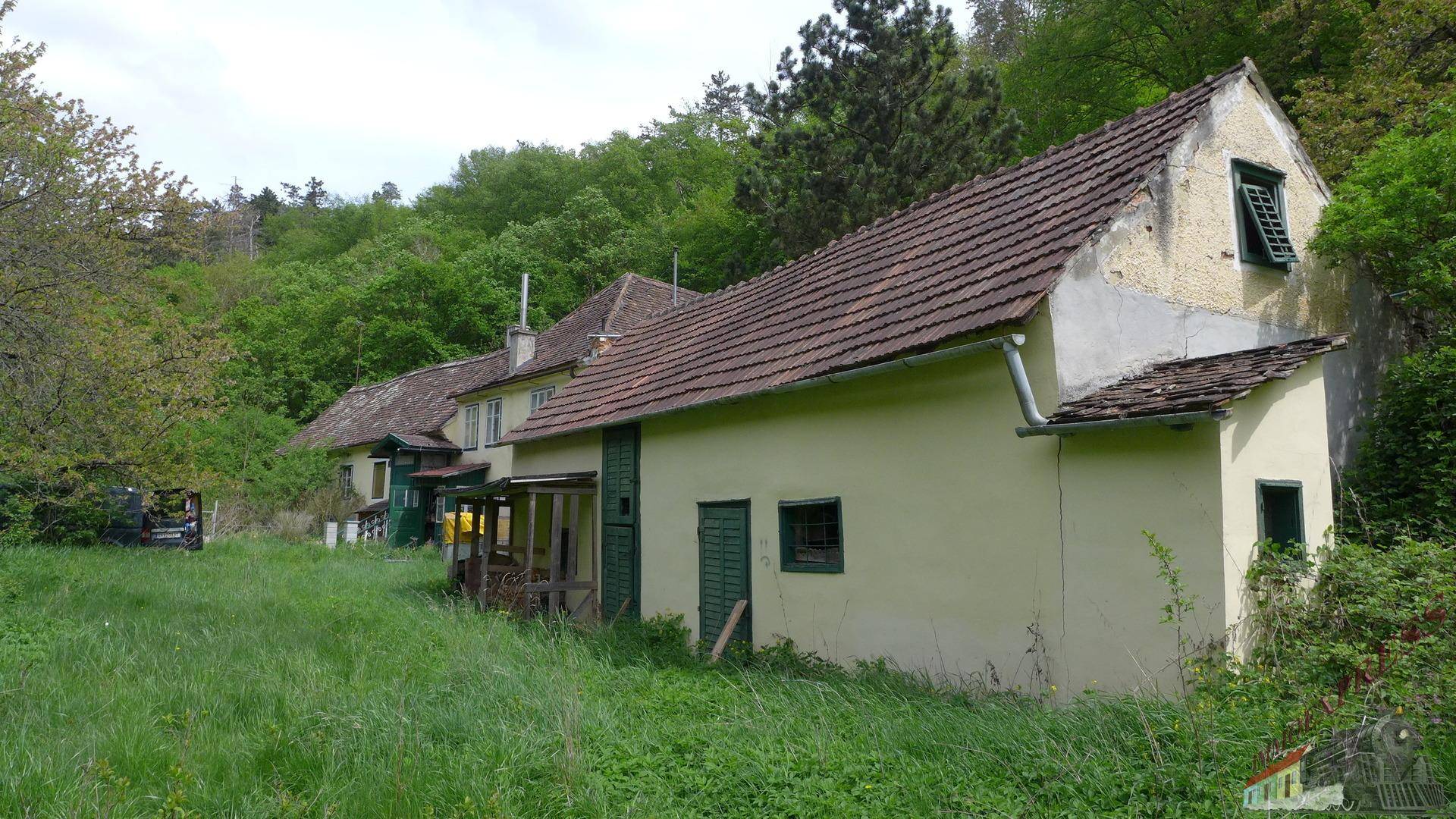 Ländliches Gebäude mit verputzter Fassade und Ziegeldach, umgeben von einer grünen Wiese und Bäumen.