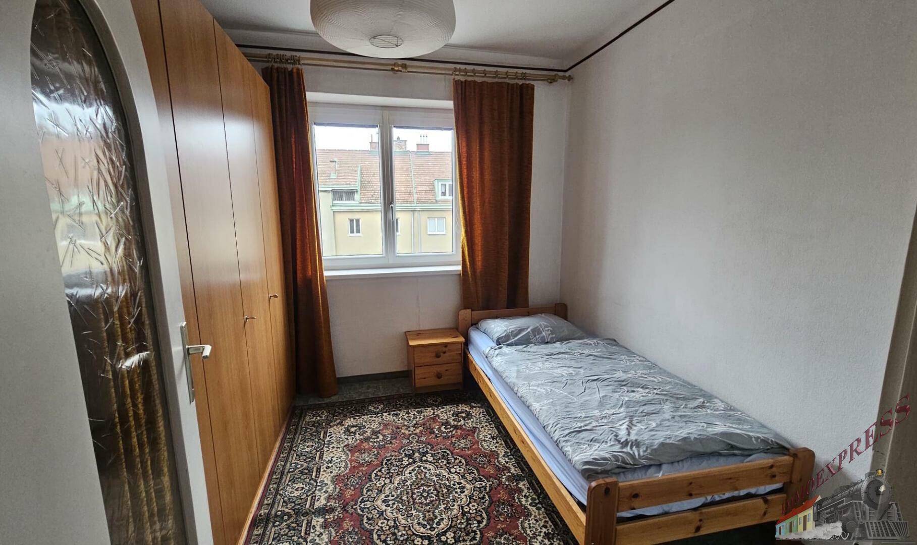 Kleines Schlafzimmer mit Einzelbett, Holzschrank und Fenster mit braunen Vorhängen.
