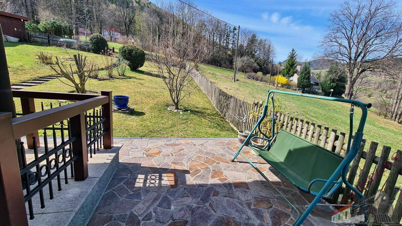 Gemütliche Terrasse mit Steinfliesen und einer Gartenschaukel, umgeben von grüner Landschaft.