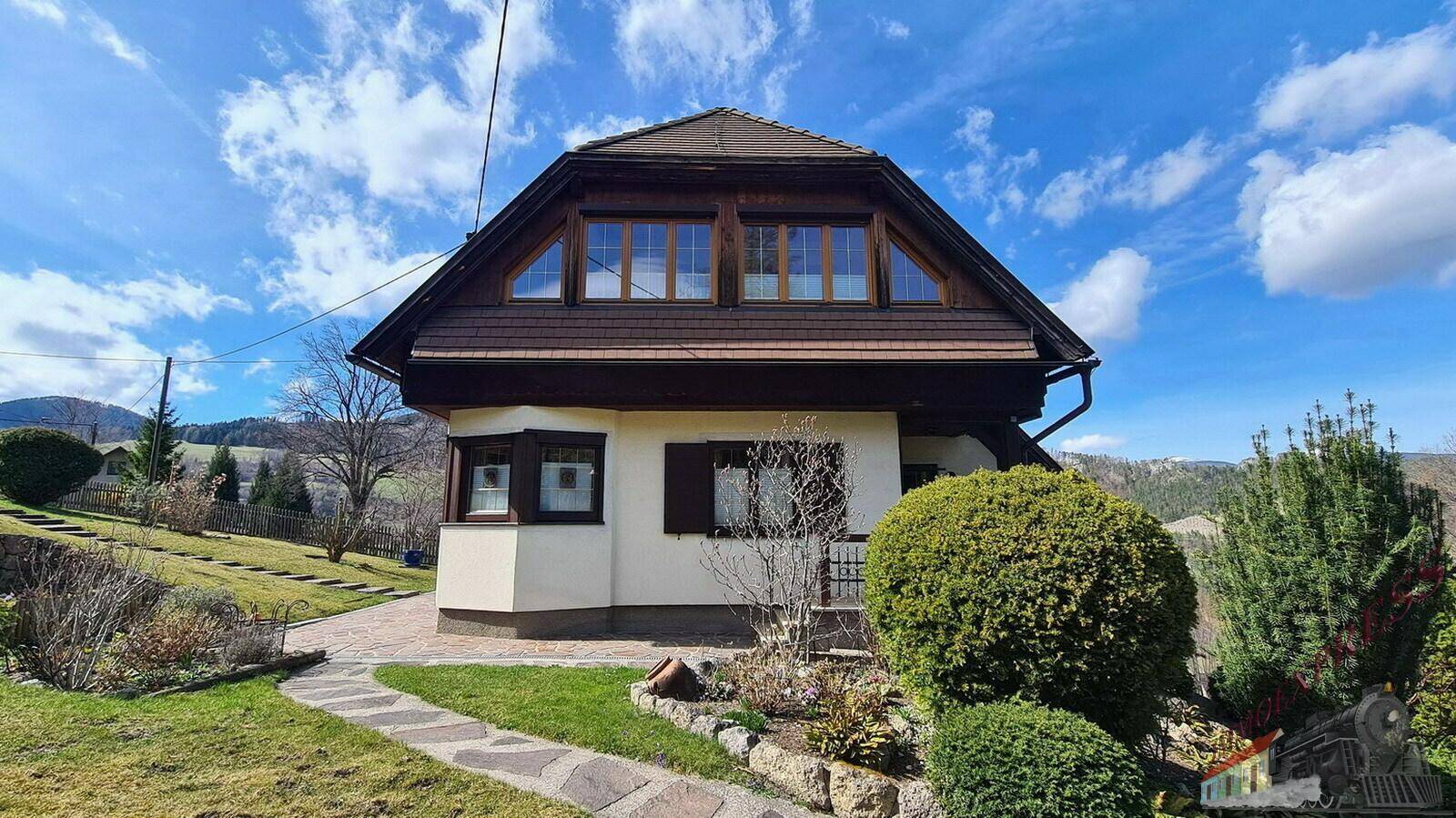 Frontansicht des Hauses mit Mansarddach und großen Fenstern, umgeben von gepflegtem Grün.