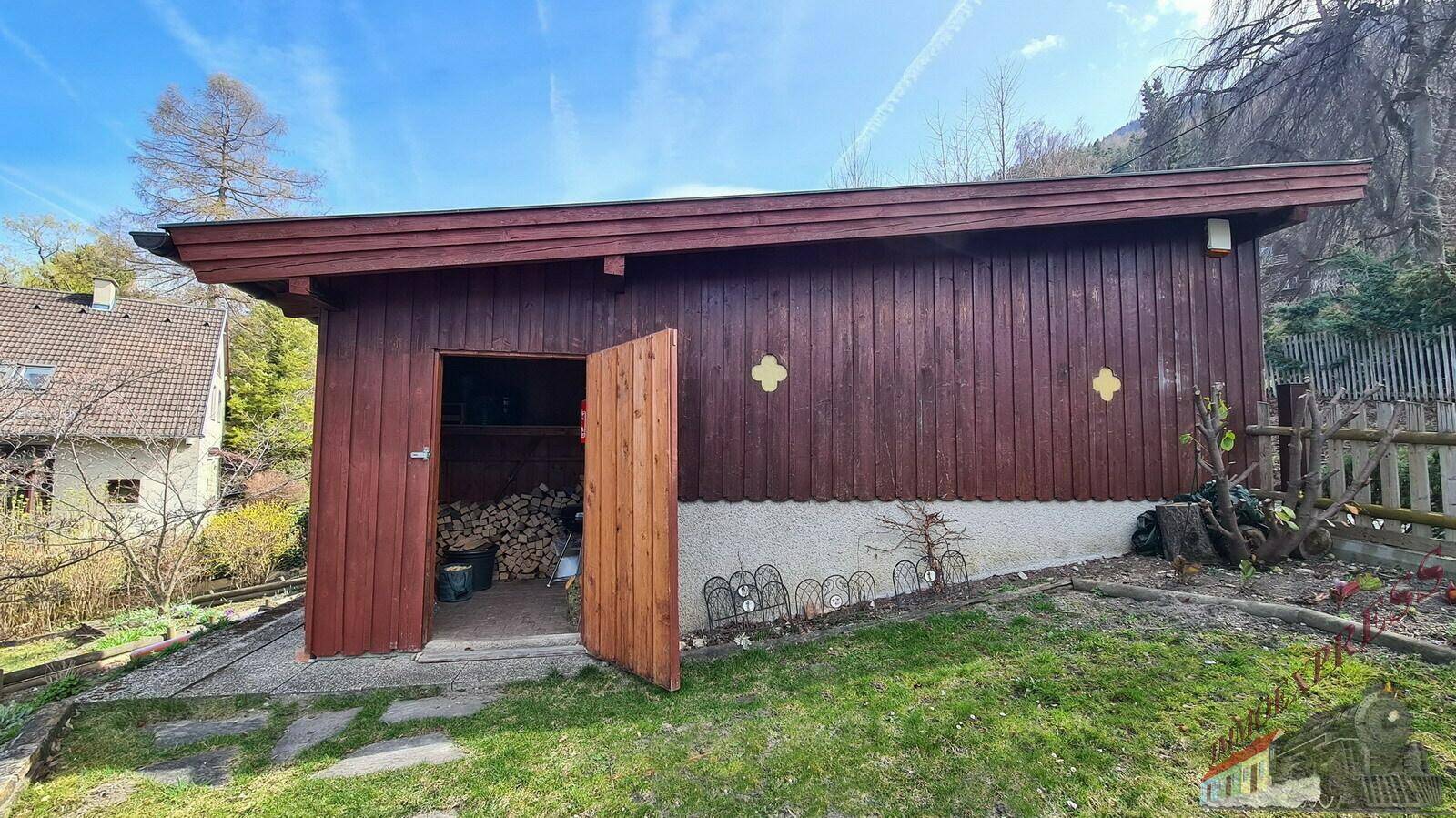 Geräteschuppen aus Holz im Garten, gefüllt mit Brennholz und umgeben von Grünflächen.