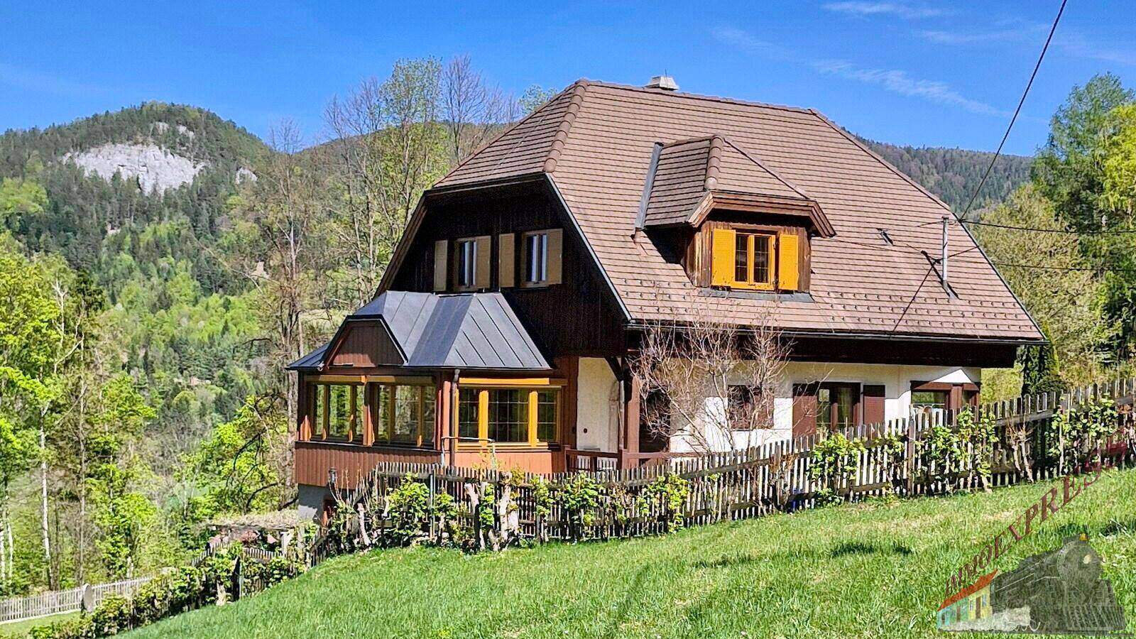 Charmantes Haus mit Mansarddach und Holzzaun, eingebettet in eine grüne Hügellandschaft.