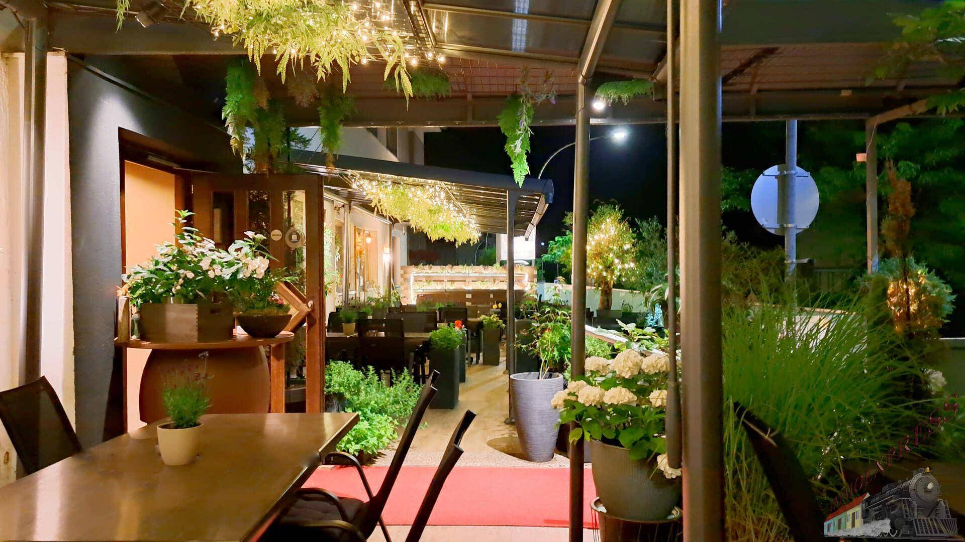 Beleuchtete Außenterrasse eines Restaurants bei Nacht, geschmückt mit Lichterketten und Pflanzen.