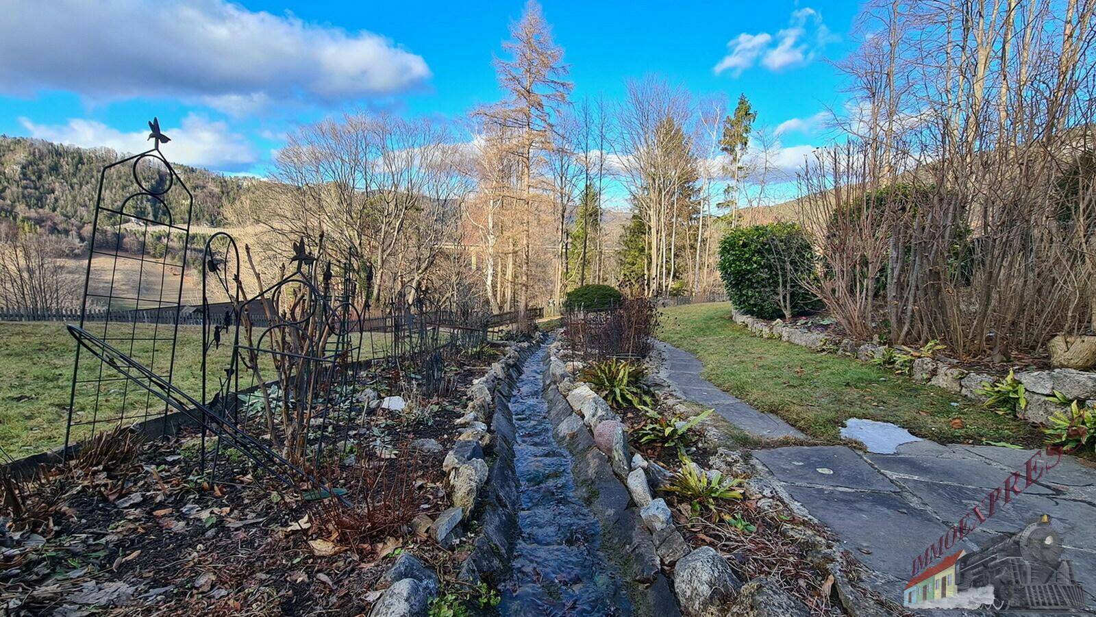 Gartenbereich mit einem kleinen Bachlauf und winterlicher Vegetation in der hügeligen Landschaft.
