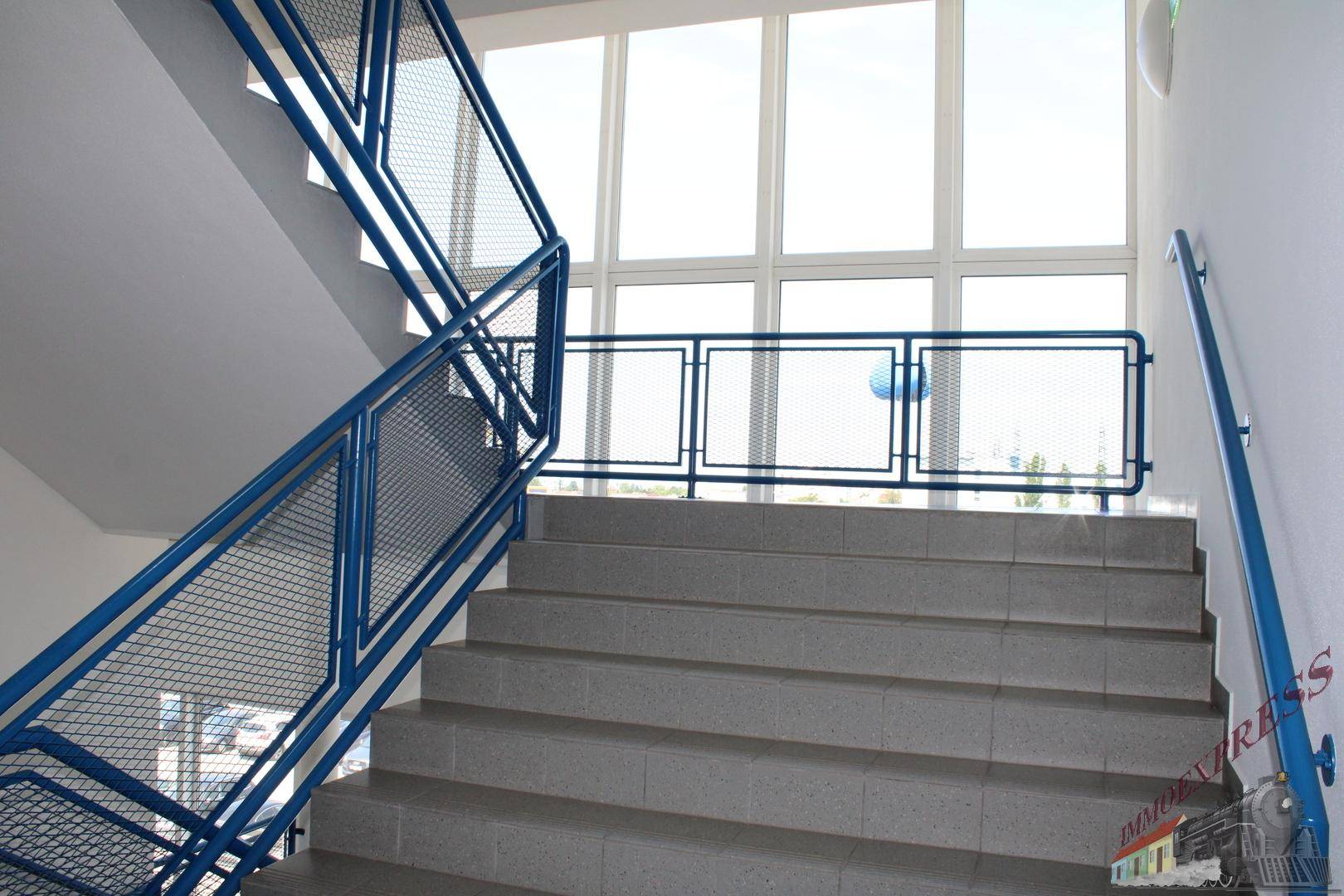 Treppenhaus mit blauen Geländern und großen Fenstern, die viel Tageslicht hereinlassen.