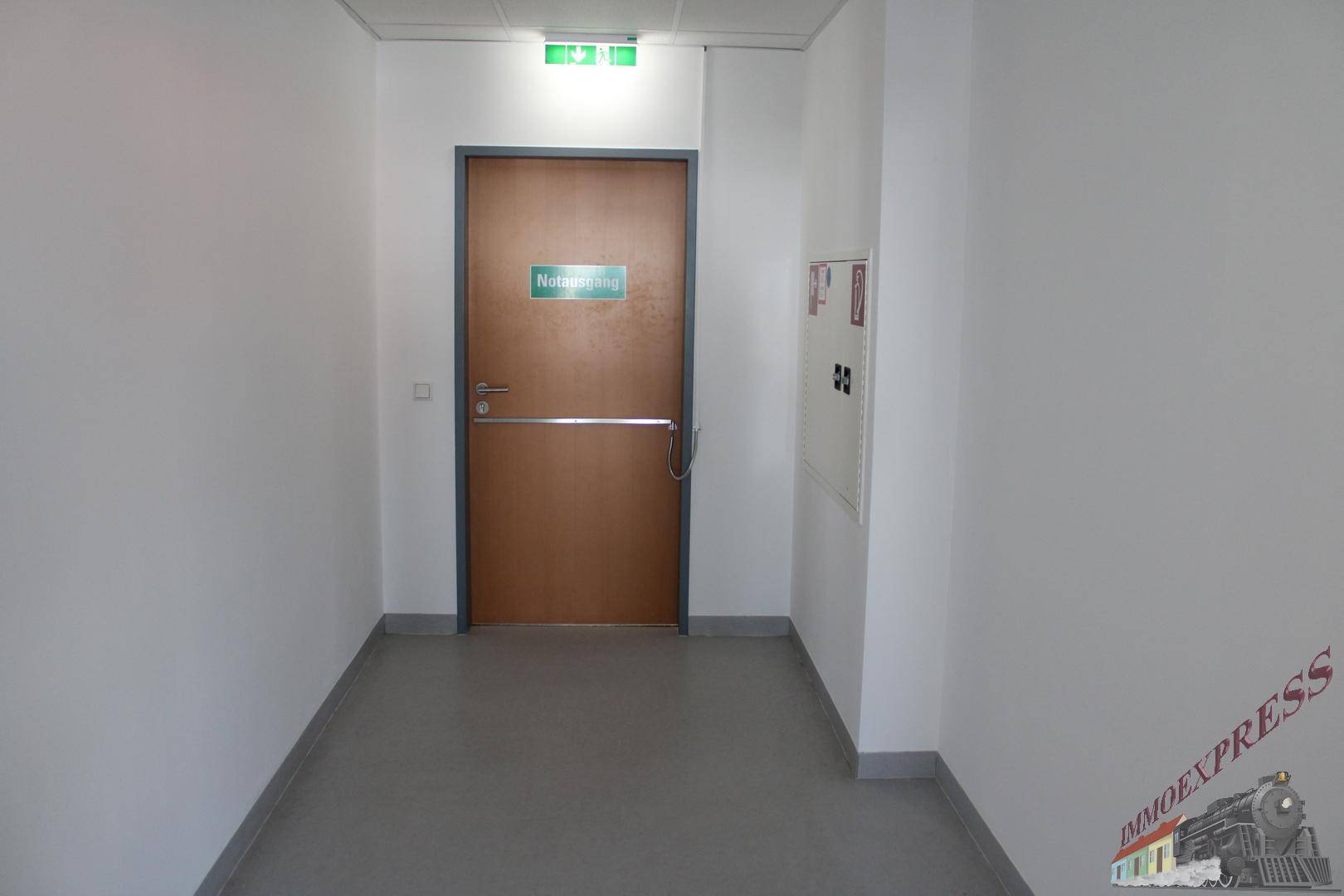 Heller Flur mit weiß gestrichenen Wänden, einer Notausgangstür und einem Feuerlöscherkasten.