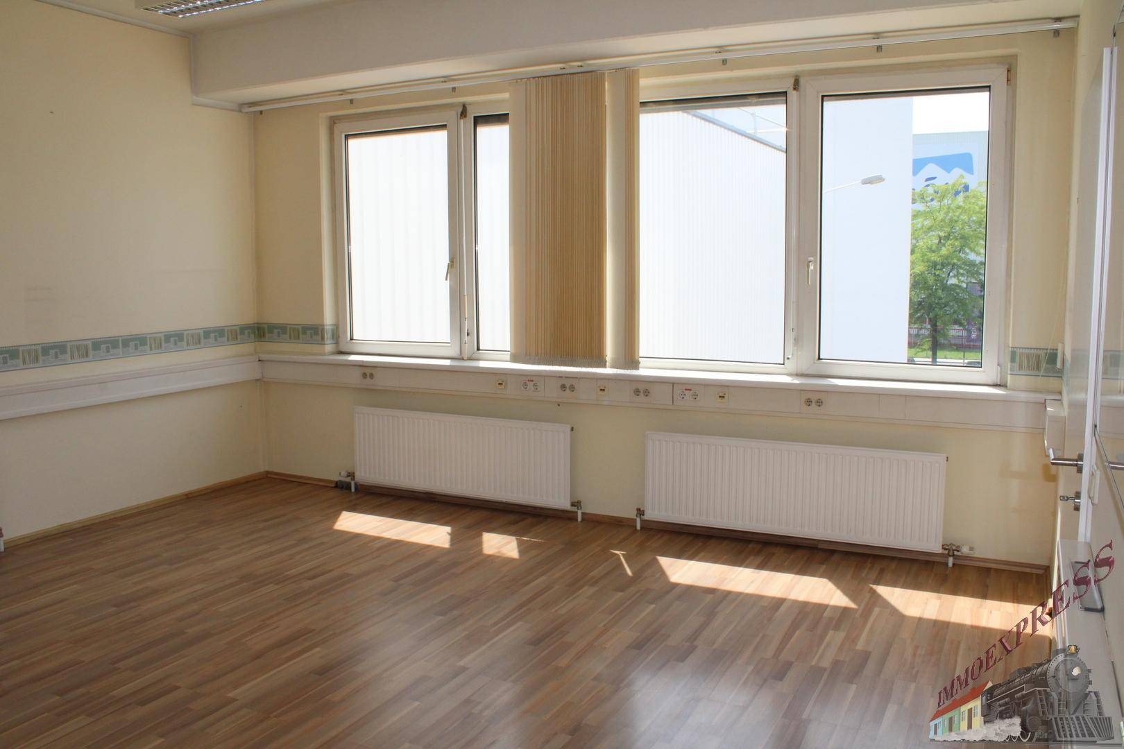Heller Büroraum mit Laminatboden, beigen Wänden und großen Fenstern.