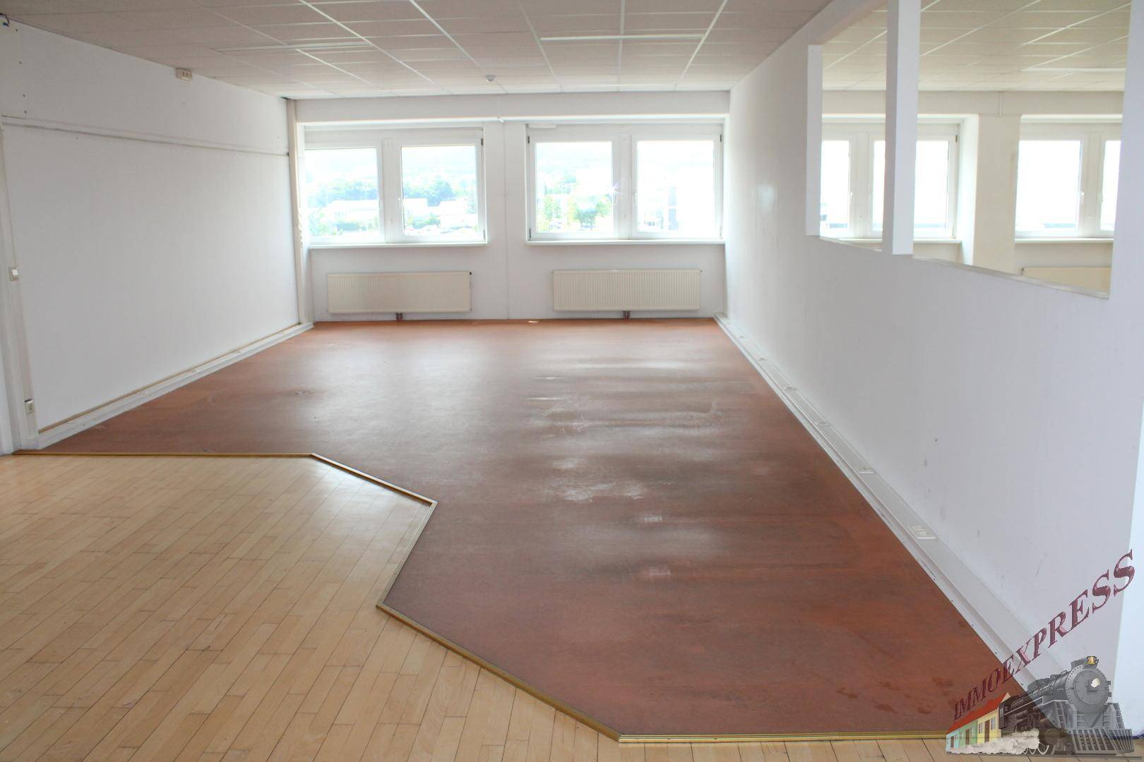 Großer Büroraum mit unterschiedlichen Bodenbelägen und natürlichem Lichteinfall durch Fenster.