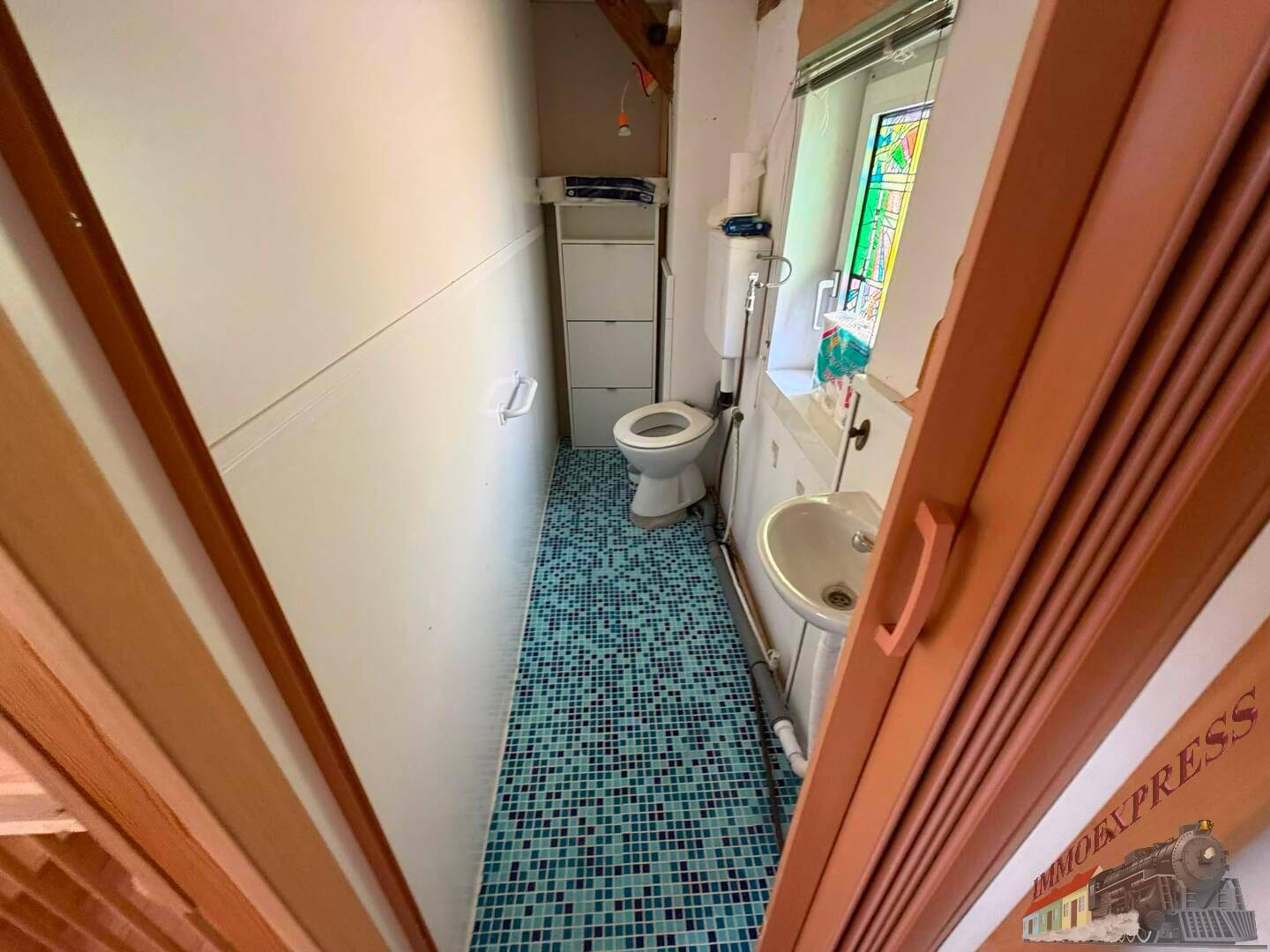 Schmale Toilette mit farbigen Mosaikfliesen, Waschbecken und Fenster.
