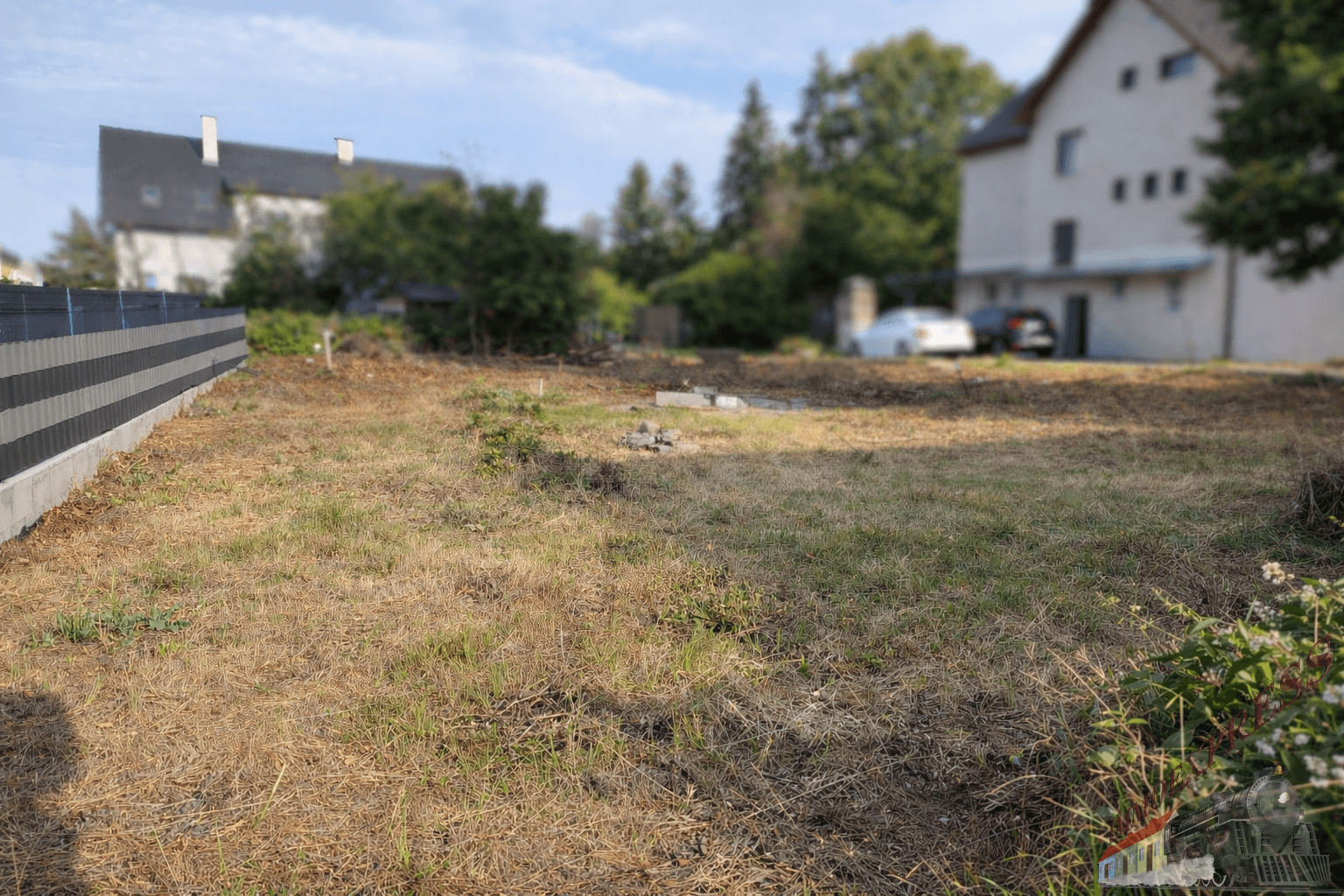 Trockenes, unbebautes Grundstück mit Zaun und unscharfen Gebäuden im Hintergrund.