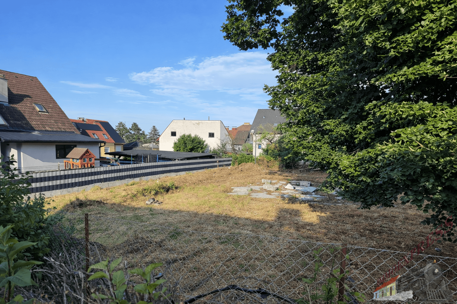 Leeres Grundstück mit trockenem Gras, umzäunt von einem Maschendrahtzaun, mit Häusern im Hintergrund.