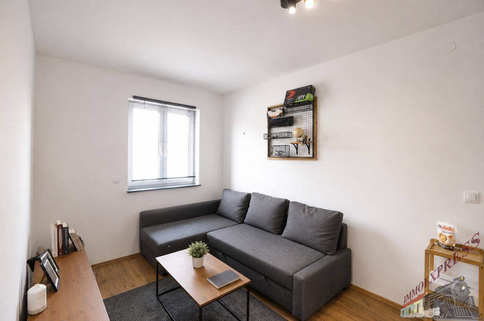 Gemütliches Zimmer mit grauem Ecksofa, Holzboden und Fenster.