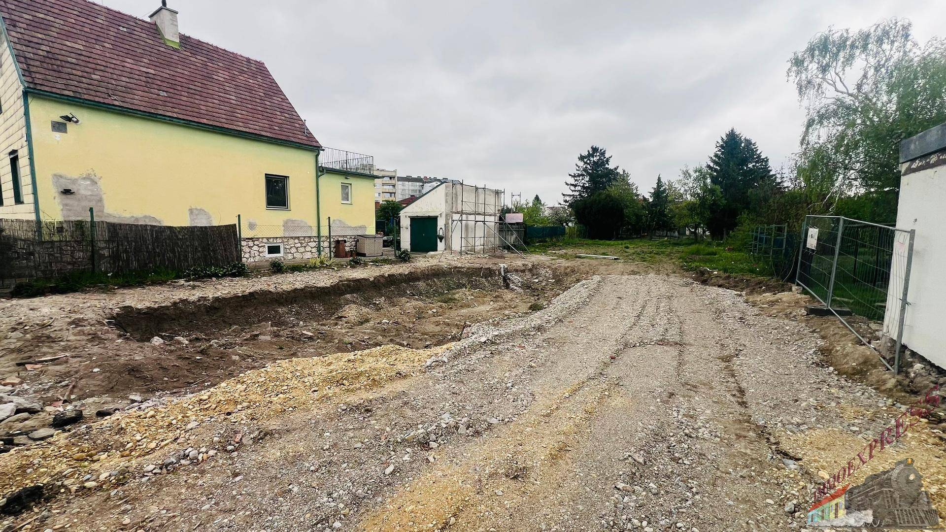 Baustelle mit ausgehobener Grube und Schotter, umgeben von Wohnhäusern.