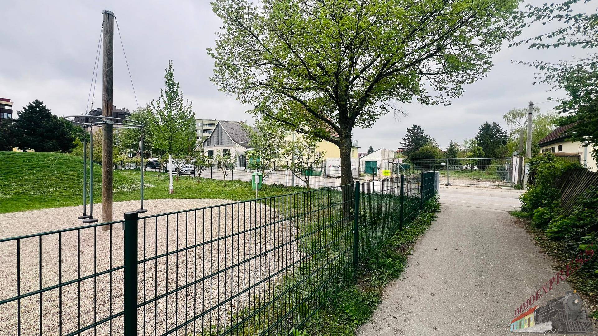 Öffentlicher Spielplatz mit Schaukeln und Bäumen, umgeben von einem Zaun.