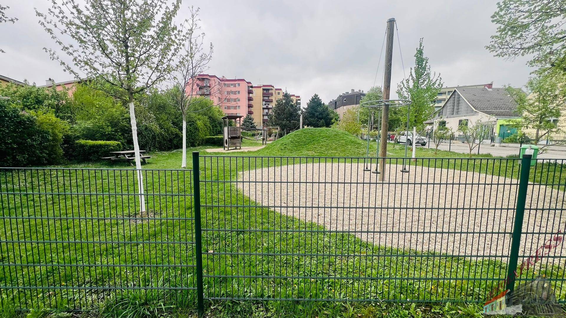Spielplatz mit Sandfläche und Schaukeln, umgeben von Grünflächen und Wohngebäuden.