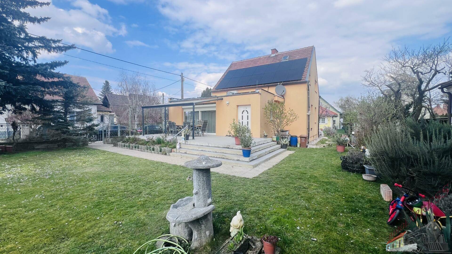 Gartenansicht mit Rasen, gepflasterter Terrasse und dem Haus mit Solaranlage.