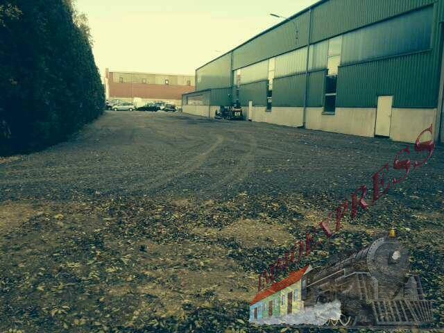 Weitläufiger Schotterplatz neben einer grünen Lagerhalle, umgeben von Bäumen und Büschen.