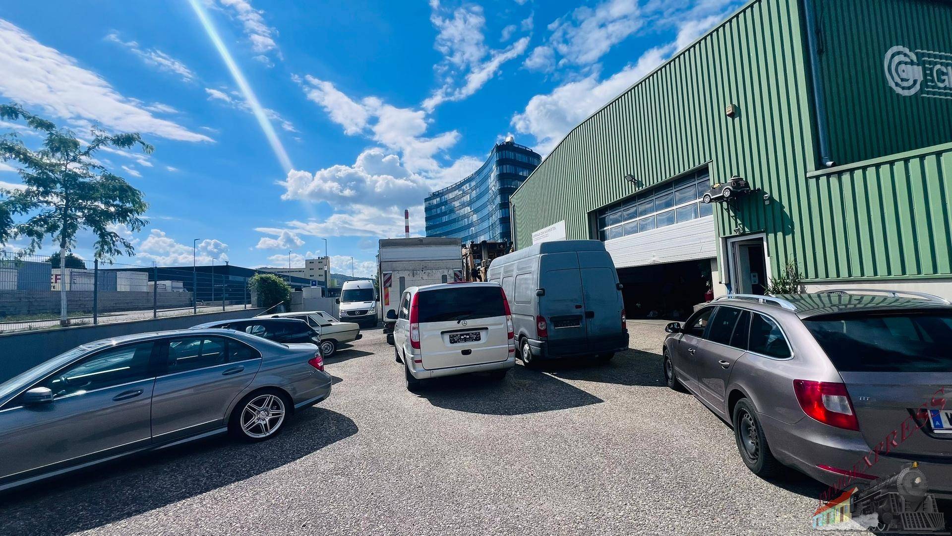Vorderansicht einer Lagerhalle mit Rolltor und geparkten Fahrzeugen, im Hintergrund moderne Bürogebäude sichtbar.