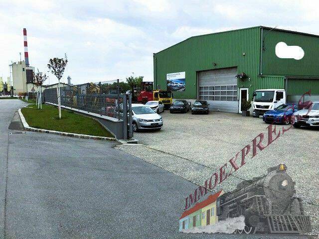 Außenansicht einer grünen Lagerhalle mit Parkplatz und einem Industriegebiet im Hintergrund, unter bewölktem Himmel.