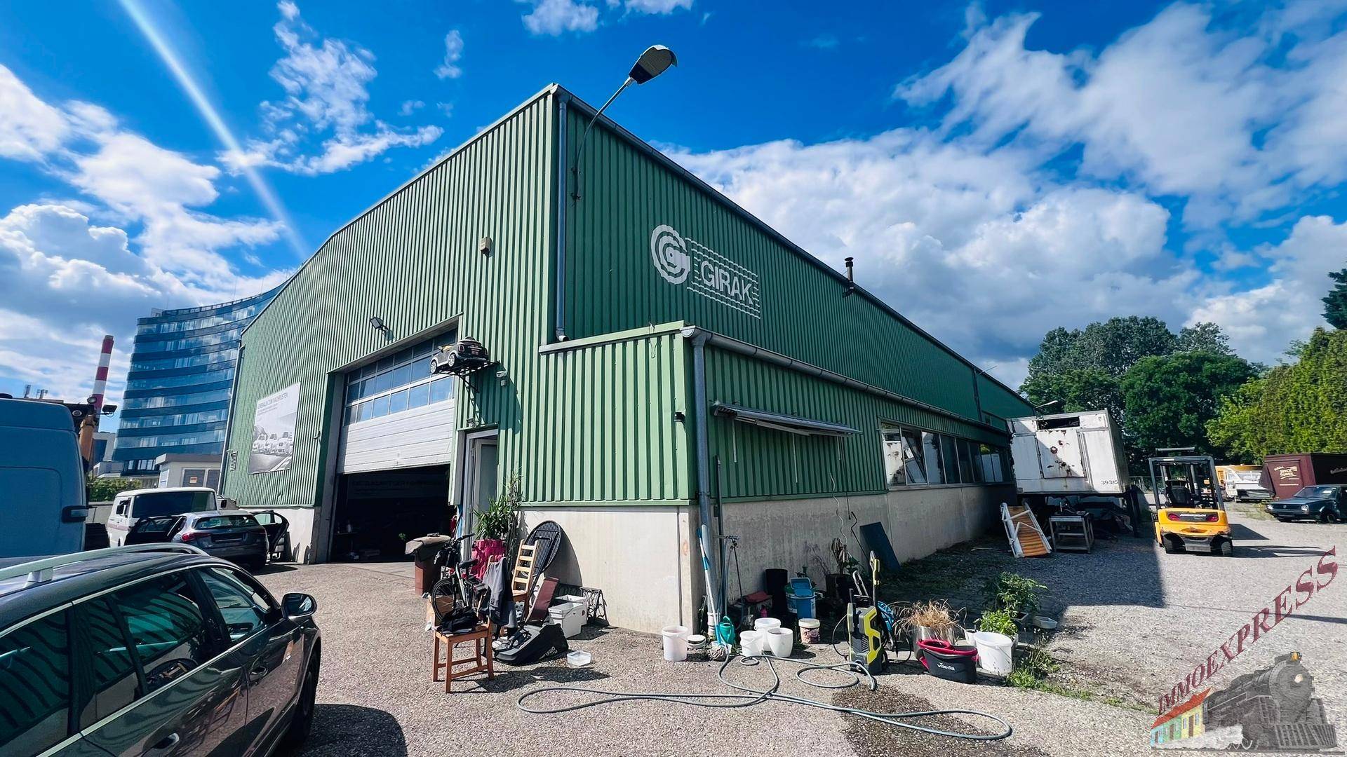 Gesamtansicht einer großen grünen Lagerhalle mit Rolltor und einem sonnigen Vorplatz mit abgestellten Gegenständen.