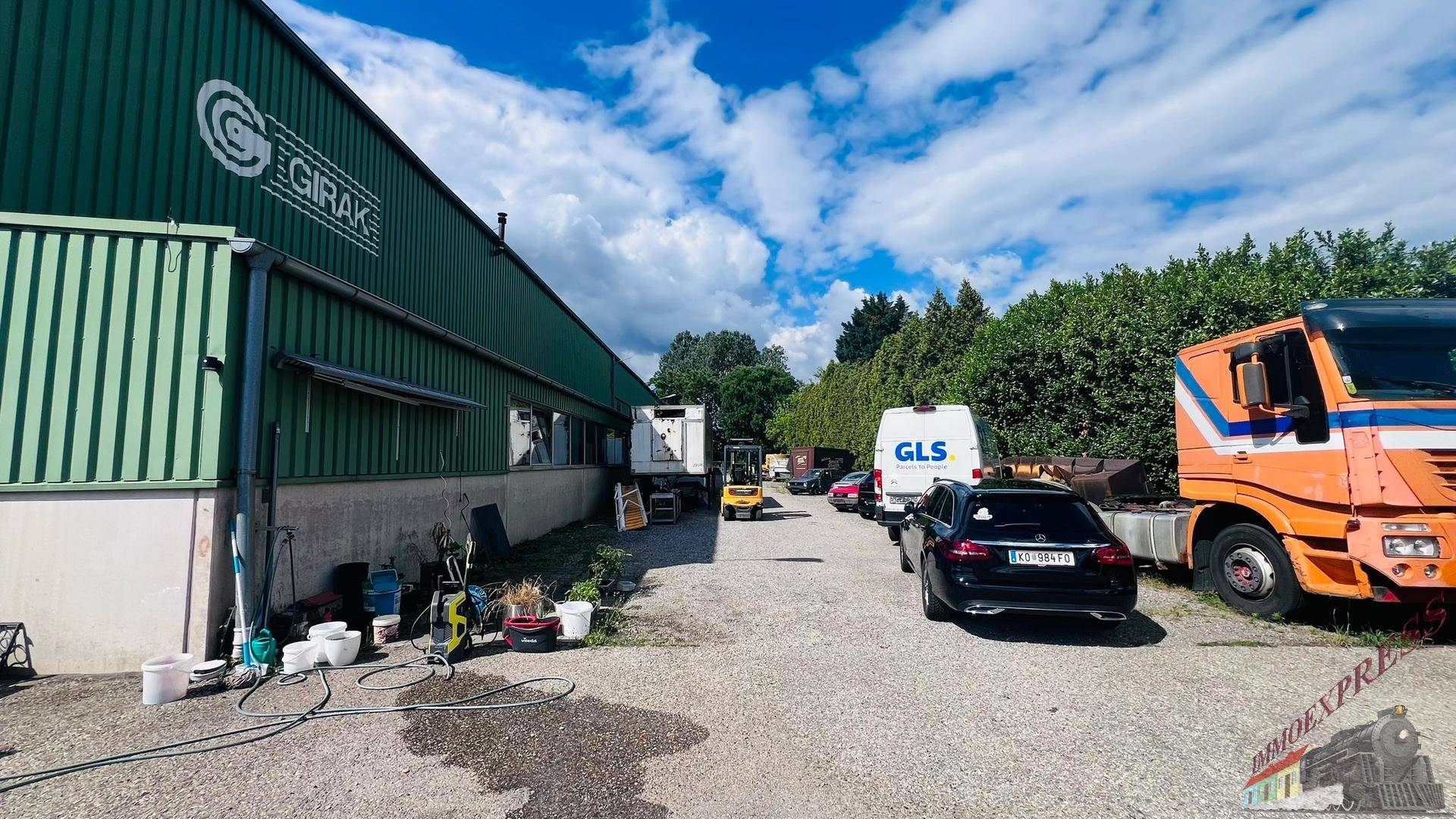 Außenbereich einer Lagerhalle mit grünem Wellblech und verschiedenen Fahrzeugen auf dem Schotterparkplatz.