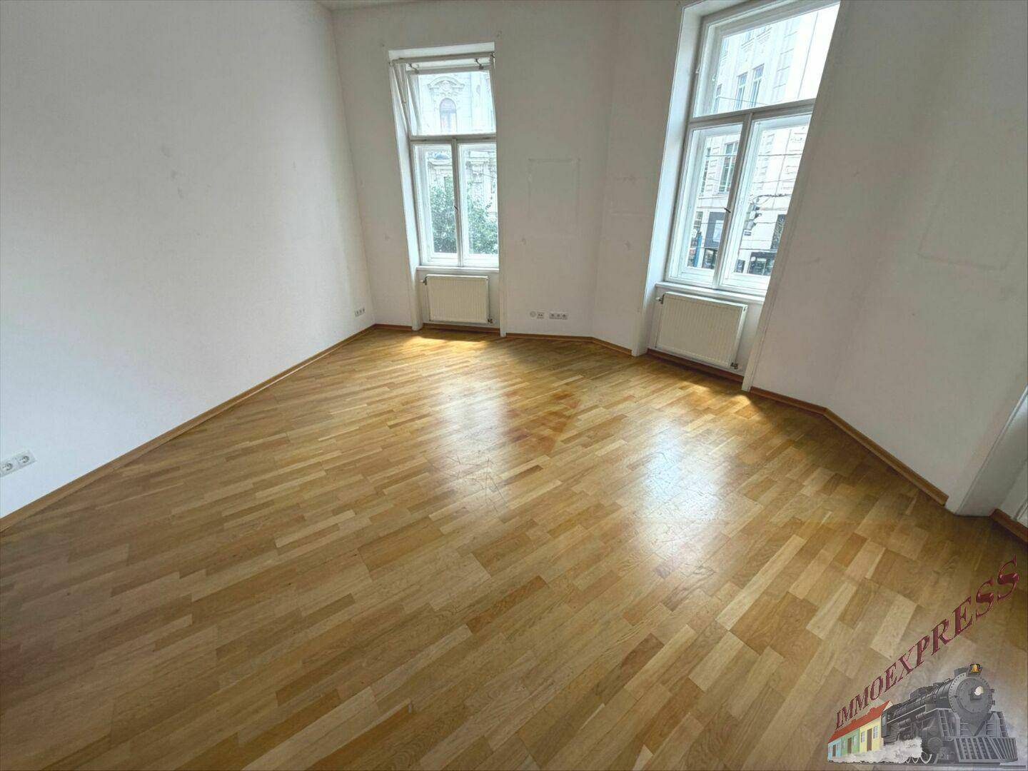 Geräumiger Raum mit hellem Holzboden, weißen Wänden und zwei hohen Fenstern zur Straße.