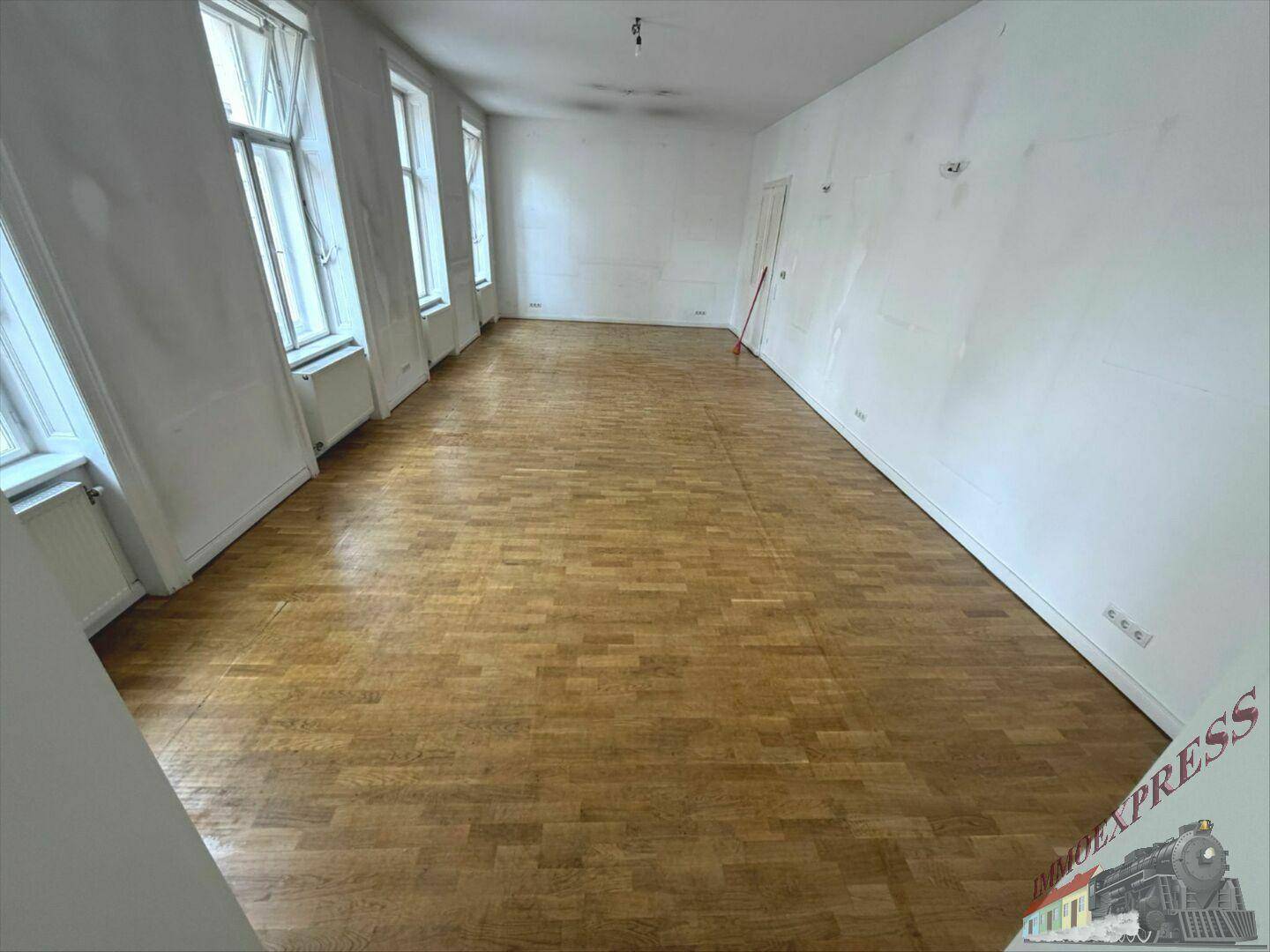 Langer, heller Raum mit Holzboden, weißen Wänden und mehreren großen Fenstern.