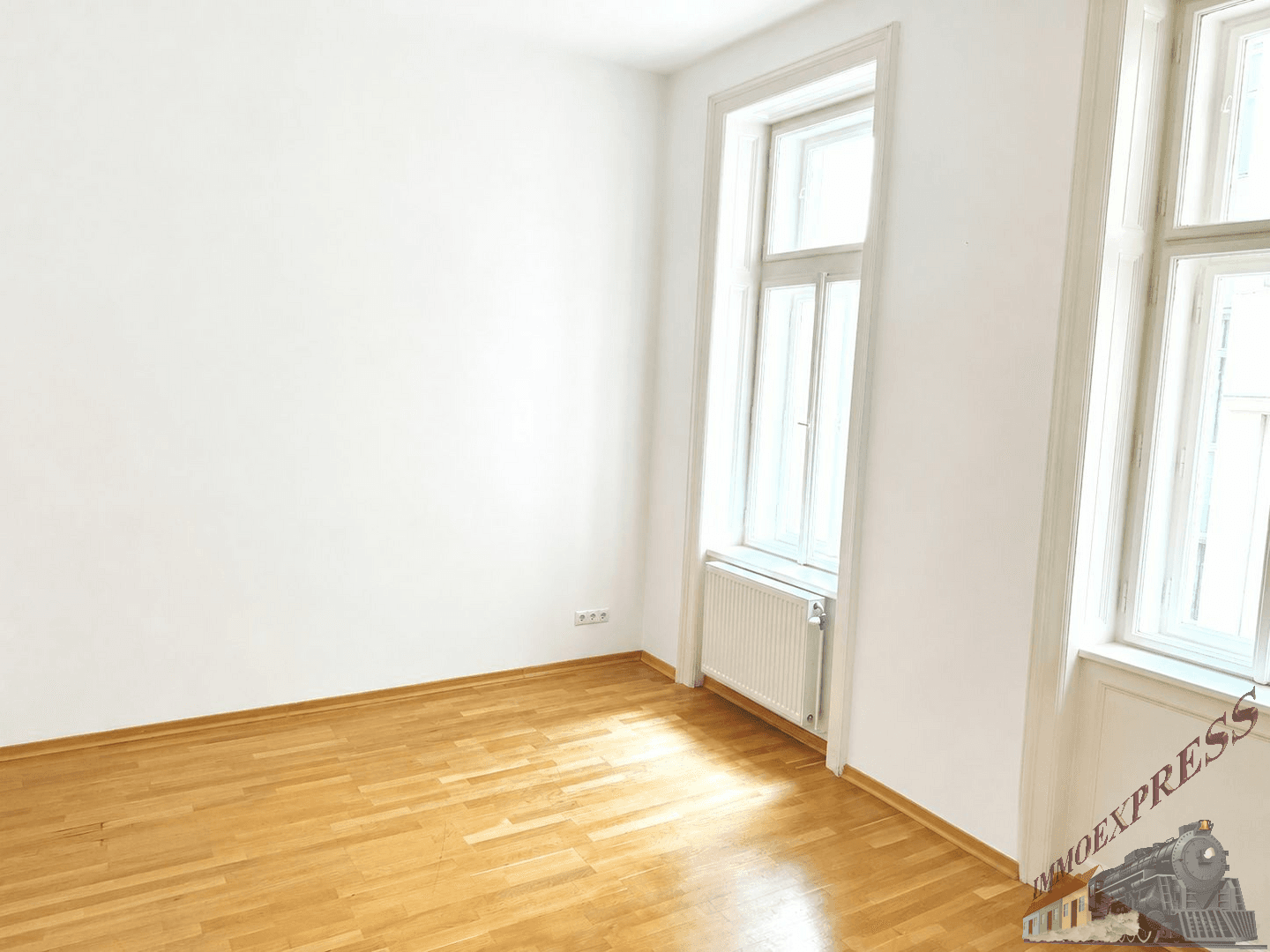 Leerer Raum mit Holzboden, weißen Wänden und zwei hohen Fenstern, die viel Licht hereinlassen.