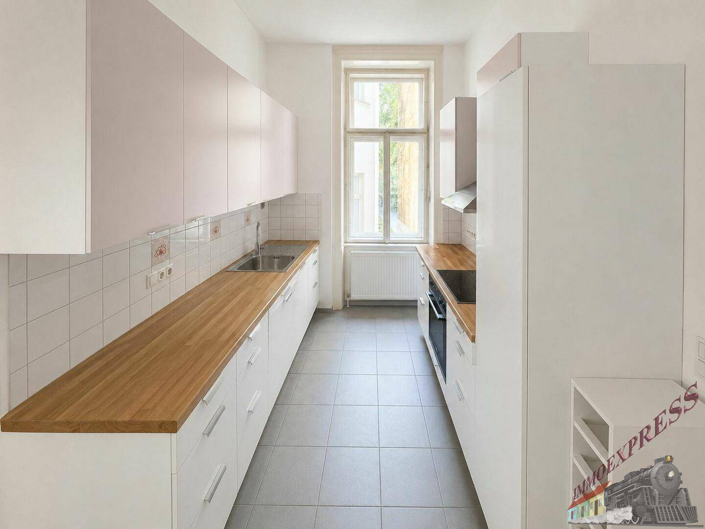 Schmale Küche mit weißen Schränken, Holzarbeitsplatten und grauem Fliesenboden sowie einem Fenster.