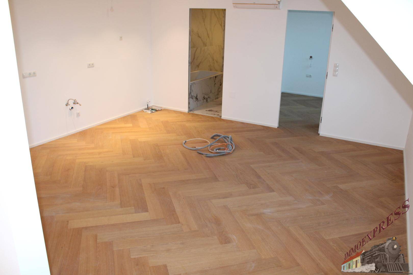 Offener Raum mit Fischgrätparkett, Verkabelung und Blick in einen gefliesten Bereich.