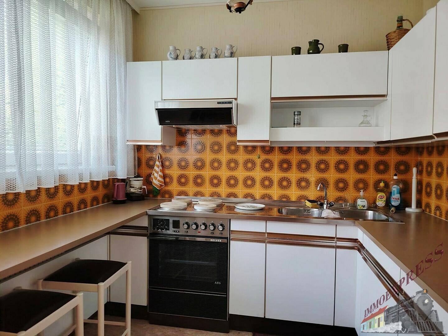 Küche im 80er-Jahre-Stil mit orangefarbenen Fliesen, weißen Schränken und Herd.