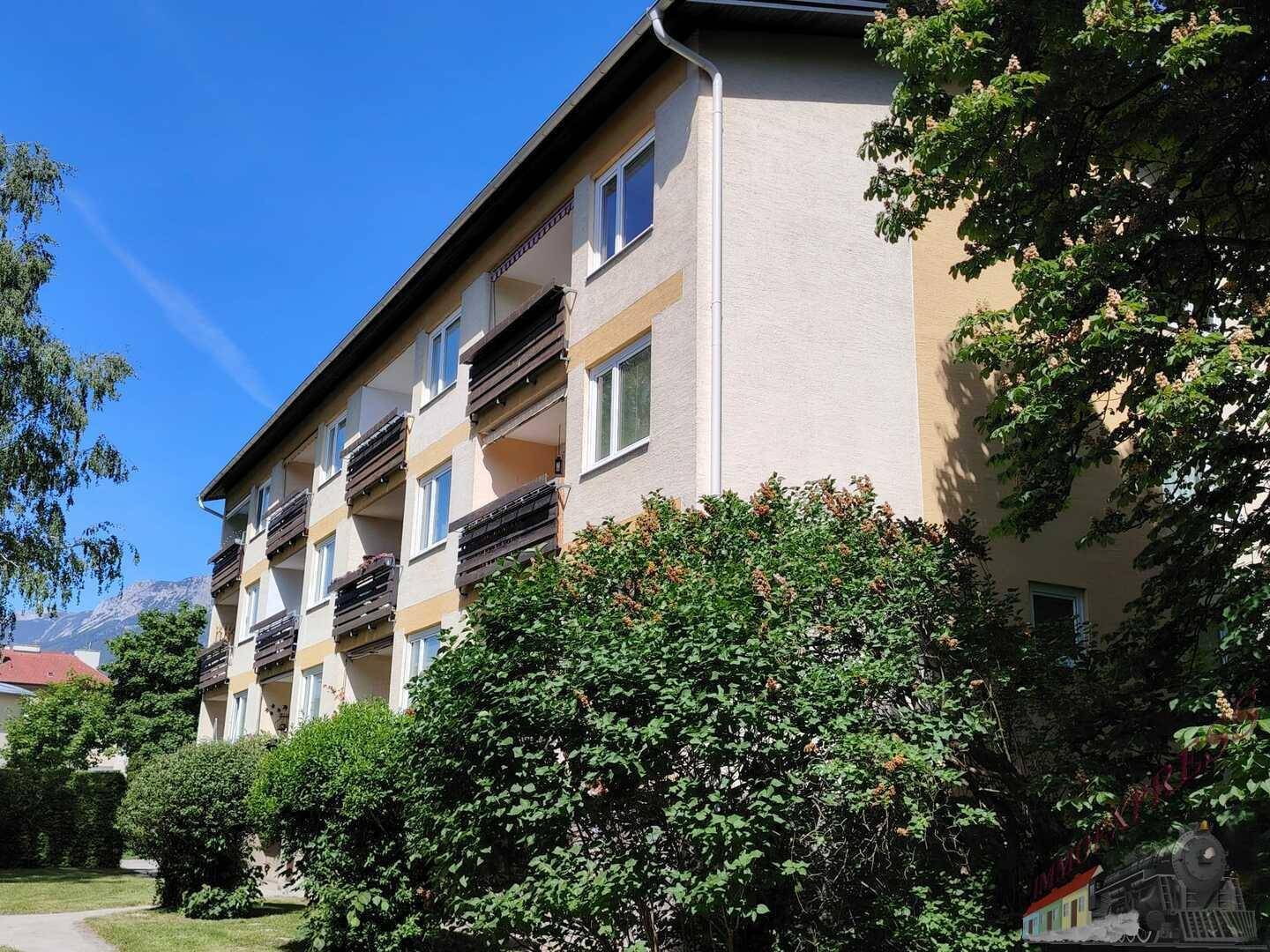 Seitenansicht des Mehrfamilienhauses mit Balkonen und Bäumen, Bergkulisse im Hintergrund.