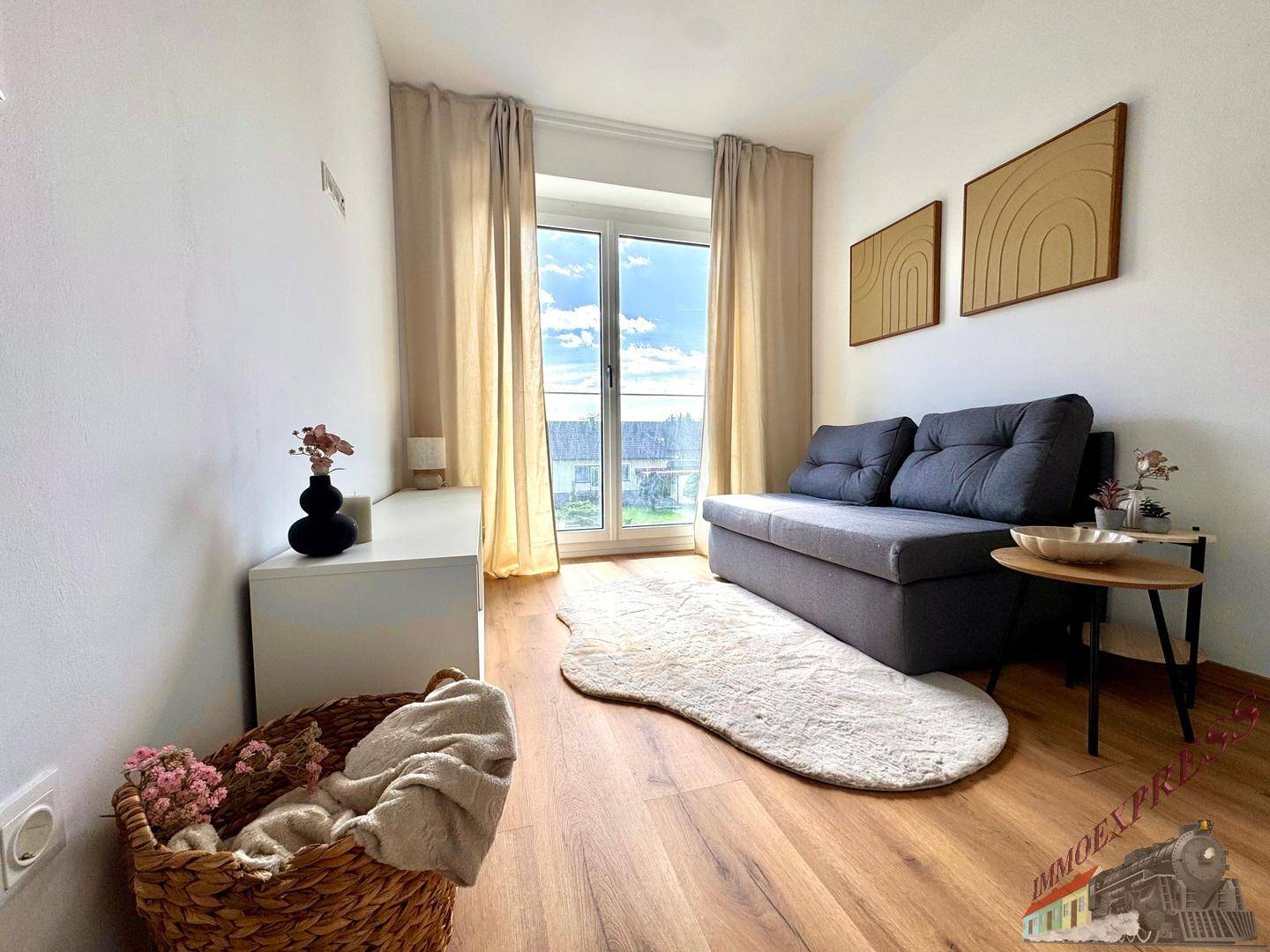 Helles Wohnzimmer mit grauem Sofa, Holzboden und großem Fenster mit Blick nach draußen.