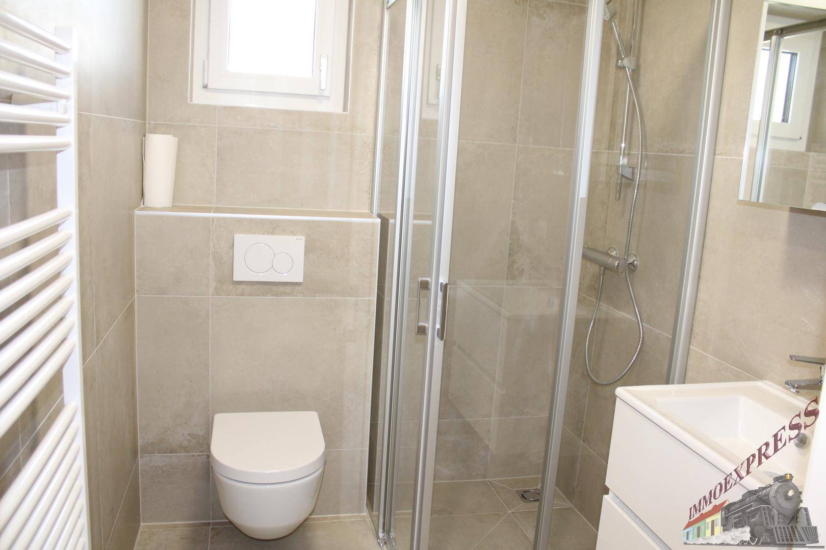 Helles Badezimmer mit Duschkabine, WC, Waschtisch, Handtuchheizkörper und Fenster für Tageslicht.