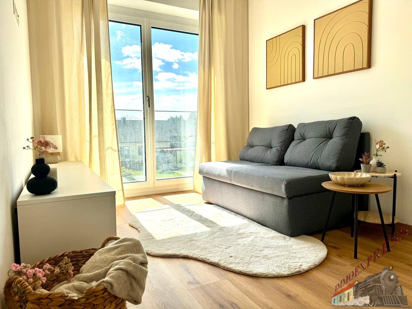 Gemütlicher Wohnbereich mit grauem Sofa, Holzboden und viel Tageslicht durch große Fenster.