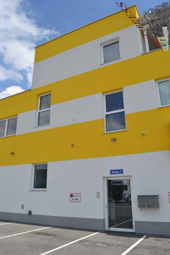 Gebäudefassade mit gelben und weißen Streifen, Eingangstür und Parkplätzen davor.
