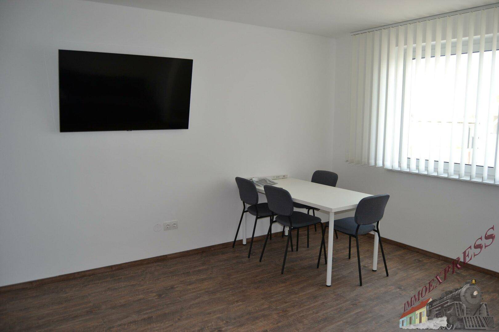 Raum mit einem Tisch, vier Stühlen und einem an der Wand montierten Fernseher.