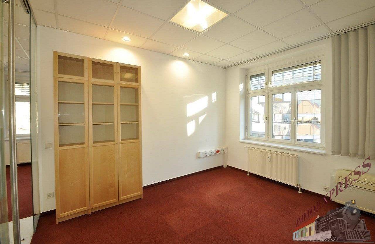 Geräumiges Büro mit Holzschrank, zwei Fenstern für viel Tageslicht und einem roten Teppichboden.