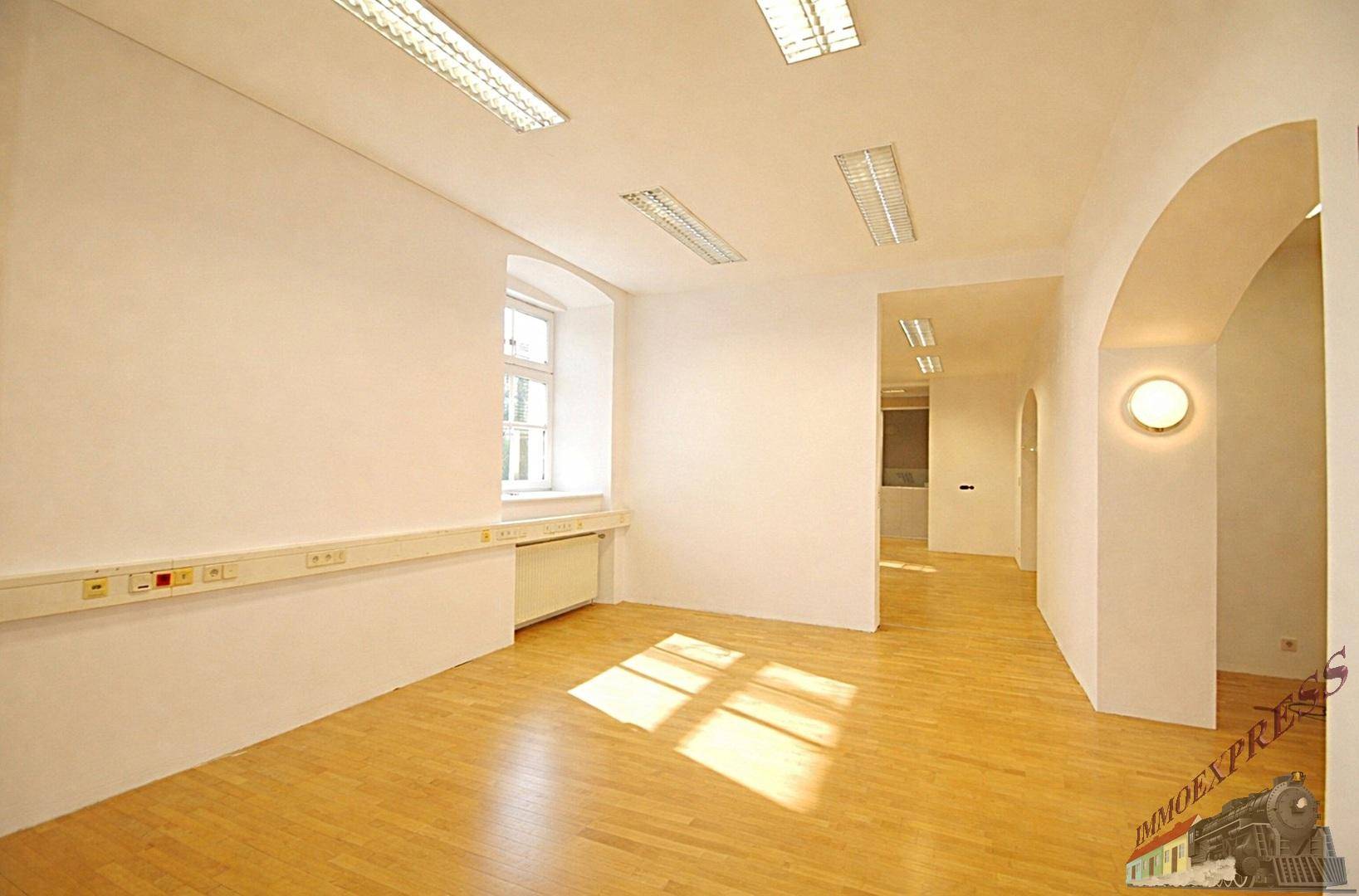 Geräumiger Büroraum mit hellem Holzparkett, großen Fenstern und einem eleganten Rundbogen.