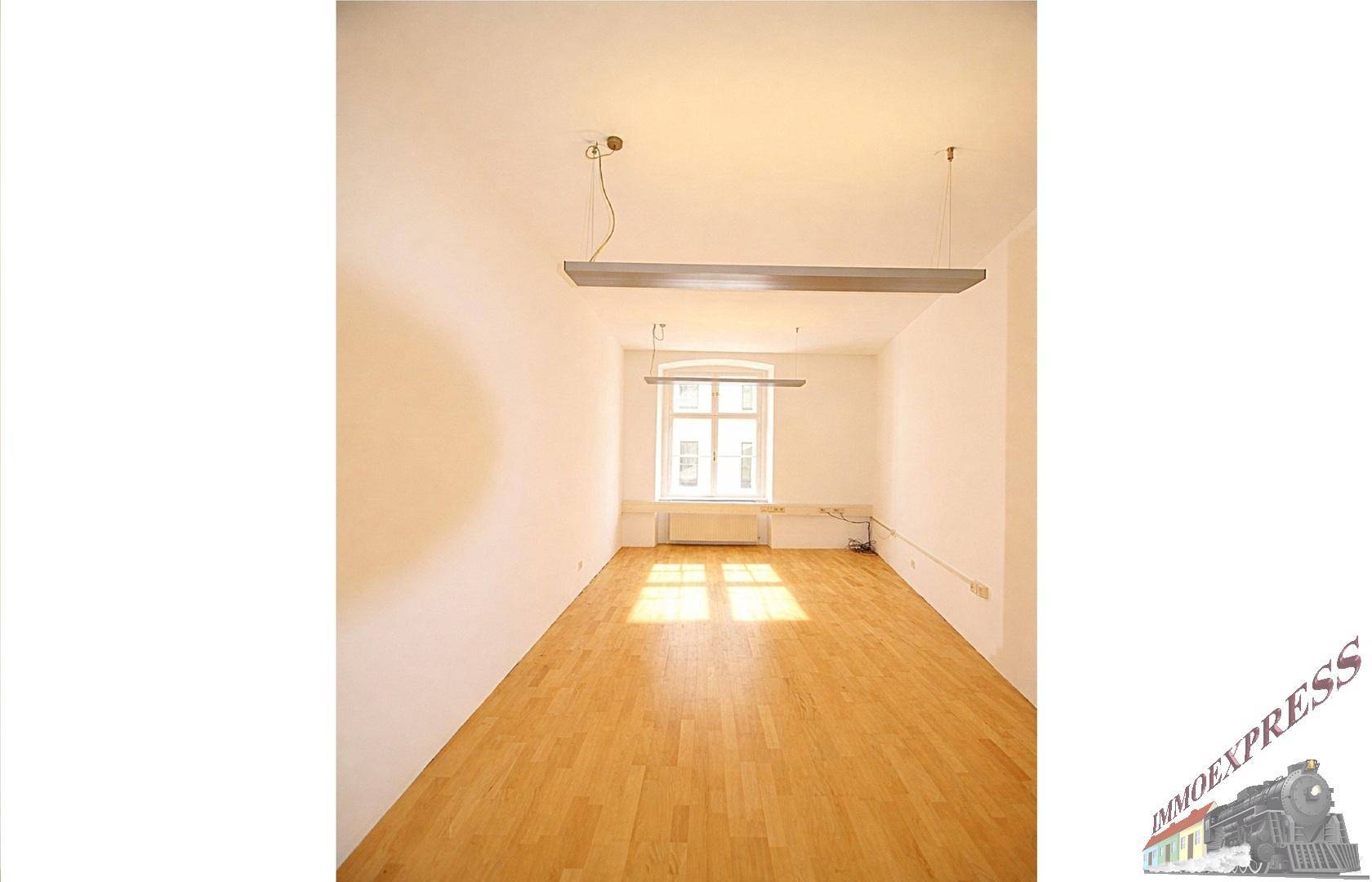 Langer, heller Büroraum mit Holzboden, einem großen Fenster und moderner Deckenbeleuchtung.