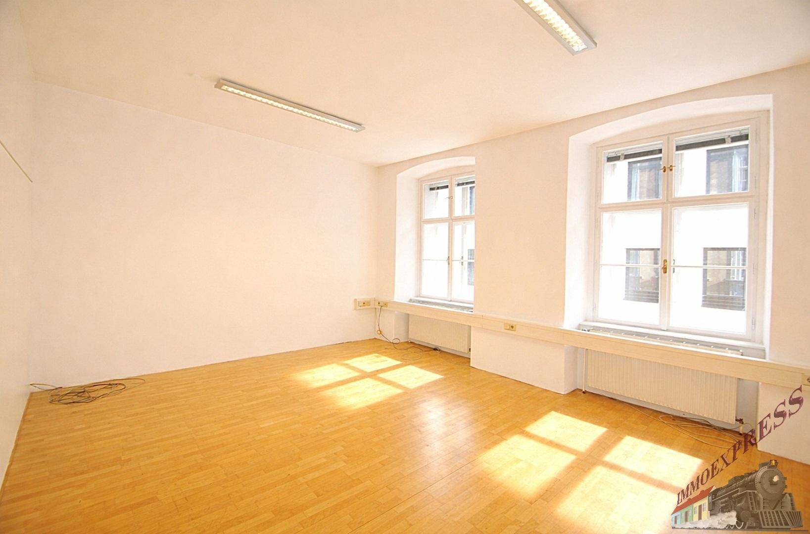 Heller Büroraum mit zwei großen Fenstern, die viel Tageslicht hereinlassen, und schönem Holzparkett.