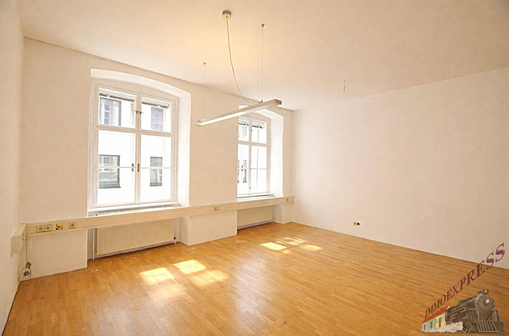 Großer, leerer Büroraum mit zwei Fenstern, die für viel Helligkeit sorgen, und robustem Holzparkett.