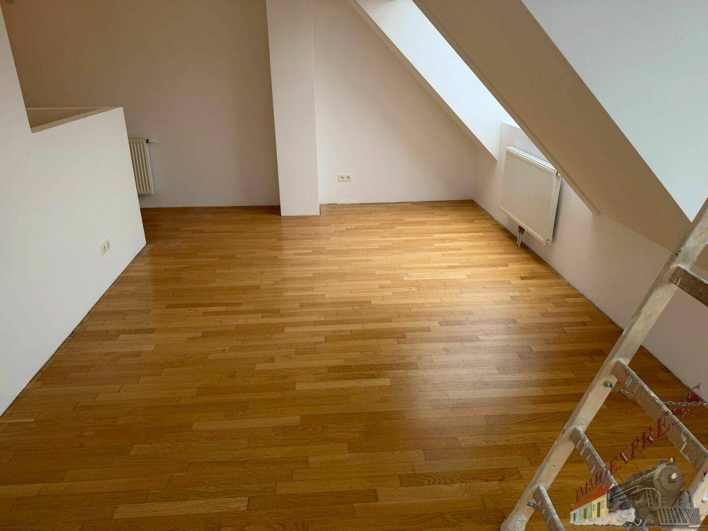 Geräumiger Dachgeschossraum mit warmem Holzboden, weißen Wänden und Heizkörper unter dem Dachfenster.
