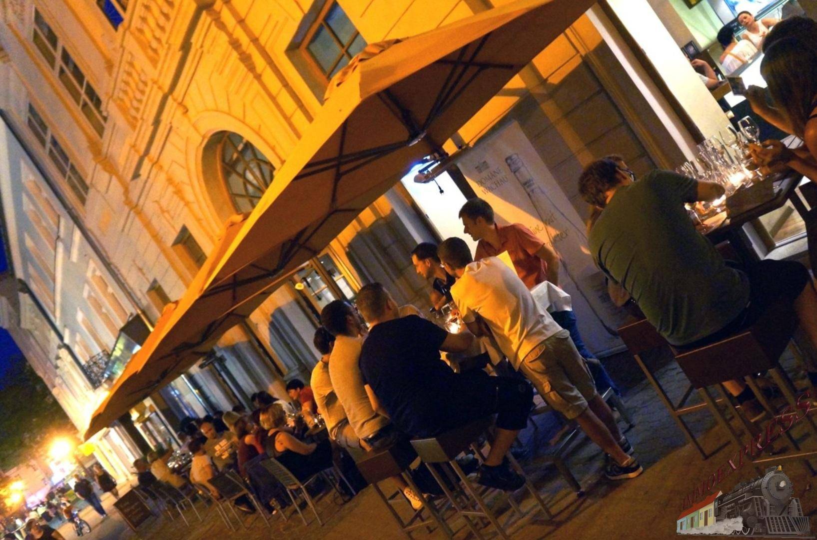 Belebte Außenterrasse am Abend mit Gästen, Tischen, Sonnenschirmen und beleuchteter historischer Fassade.