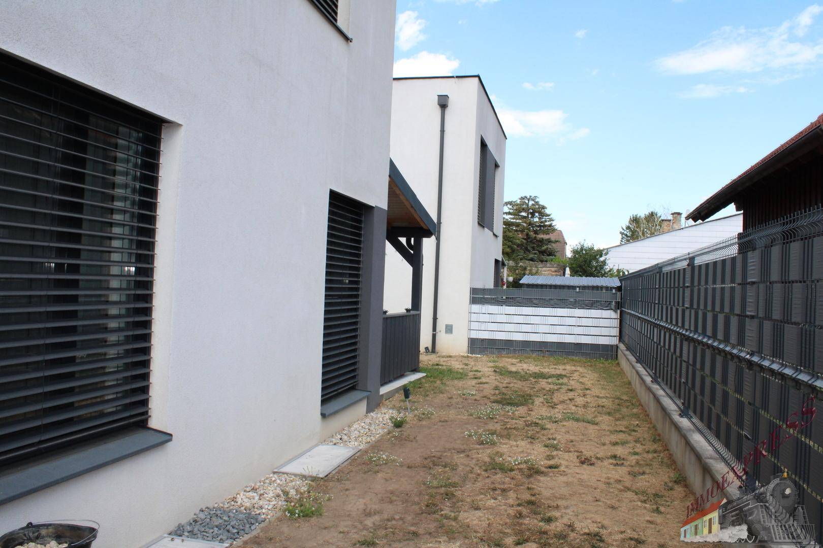 Seitenansicht des Hauses mit kleinem Gartenbereich und Zaun, umgeben von Grün.