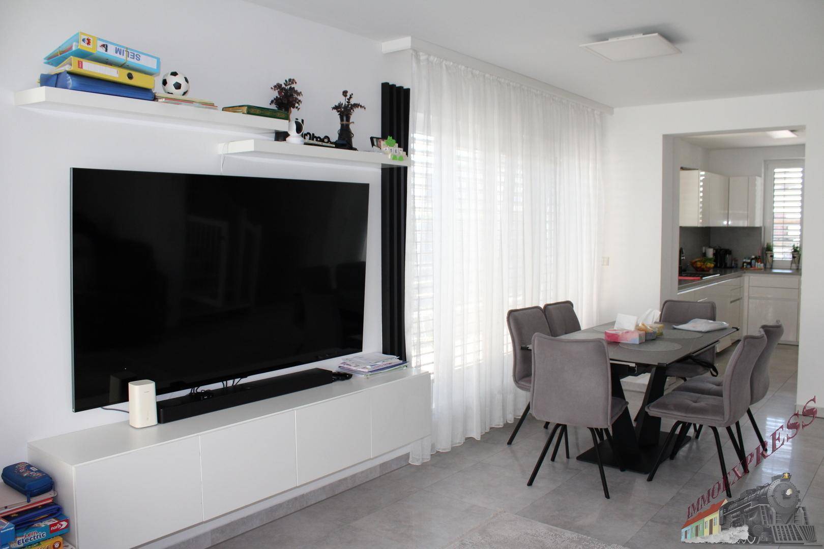 Heller Wohn- und Essbereich mit großem Fernseher, Esstisch und Blick in die angrenzende Küche.