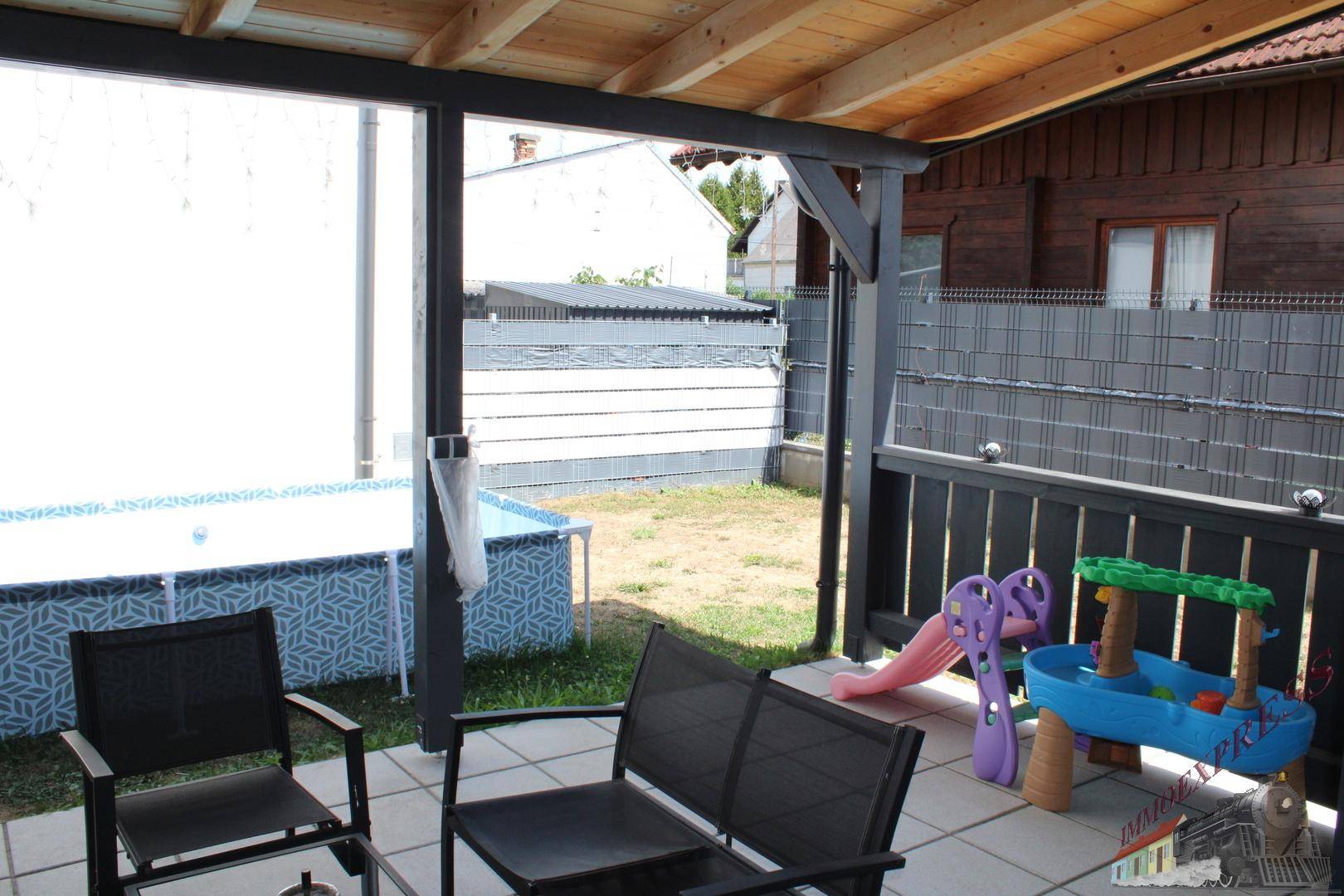 Überdachte Terrasse mit Sitzgelegenheiten, Kinderspielzeug und einem Aufstellpool im Garten.