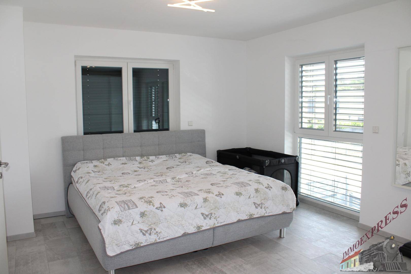 Schlafzimmer mit grauem Bett, Babybett und zwei Fenstern mit Außenjalousien für Privatsphäre.