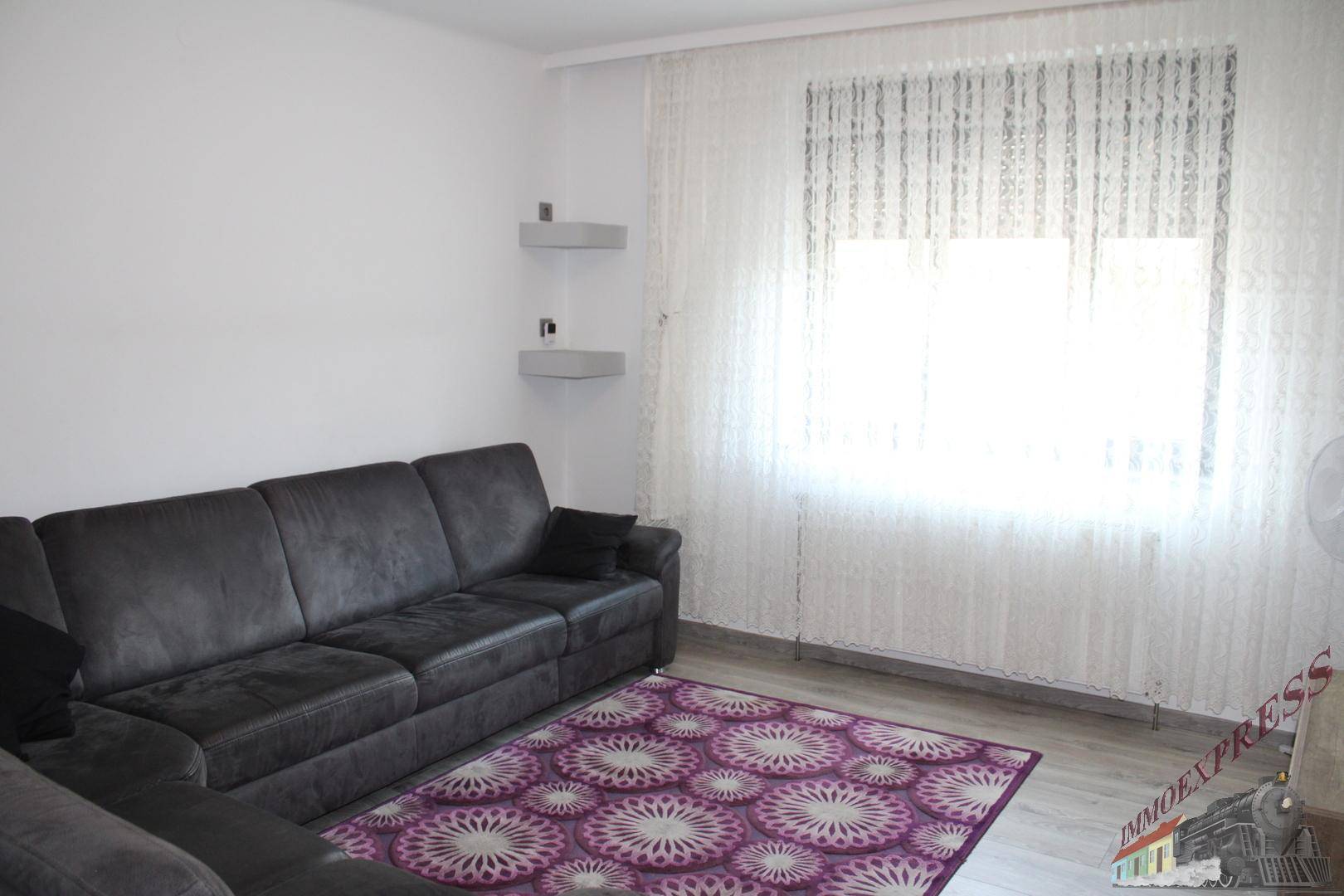 Helles Wohnzimmer mit einem großen grauen Sofa und einem gemusterten Teppich.