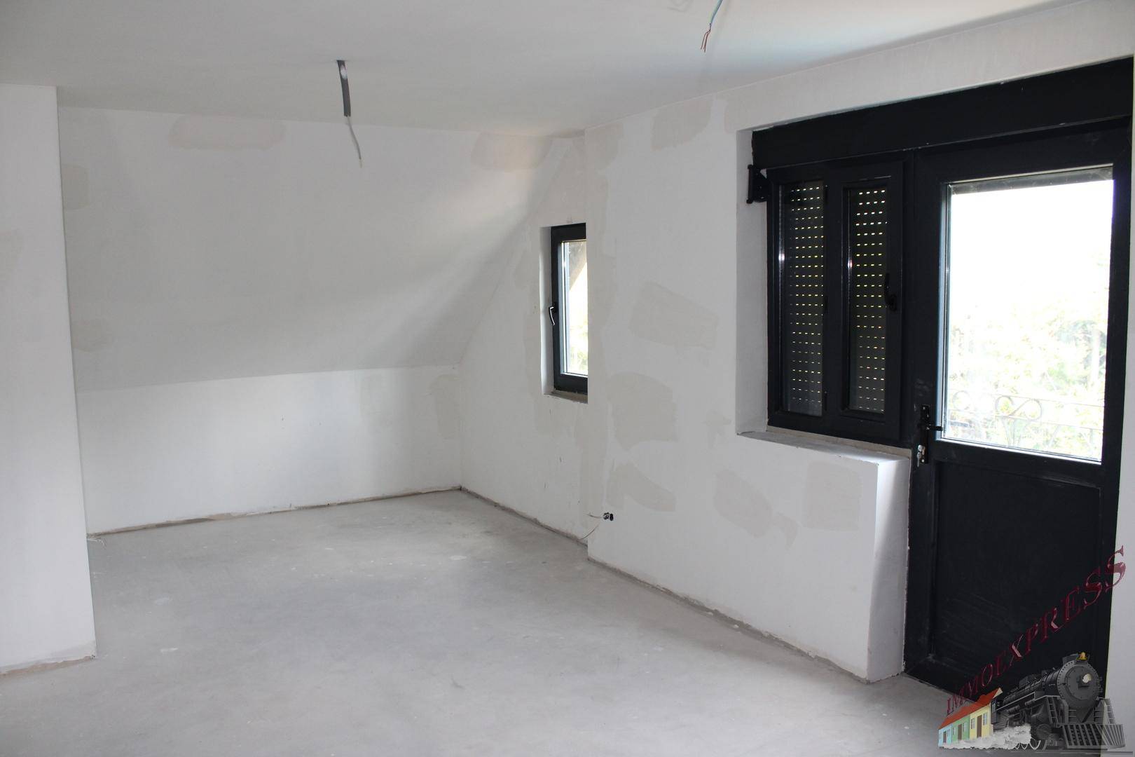 Unfertiger Raum im Dachgeschoss mit schrägen Wänden und Fenster mit Rollladen.